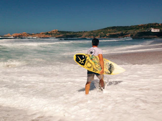 Surfer carrying a board along a rugged Baja beach with crashing waves.