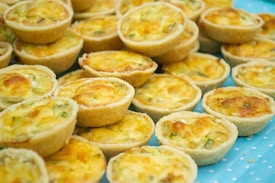 A collection of mini quiches with golden-brown tops and sprinkled with small green bits, possibly scallions or herbs. They are arranged closely together on a light blue surface with white polka dots.