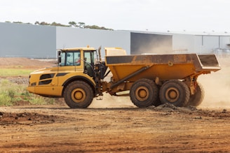 A large, yellow articulated dump truck is moving across a construction site. The truck has a spacious, open cargo area and oversized black tires. Dust is being kicked up by its movement, indicating it is actively being used in a dirt-laden environment. In the background, an industrial building with a flat roof stands near the site.