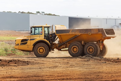 A large, yellow articulated dump truck is moving across a construction site. The truck has a spacious, open cargo area and oversized black tires. Dust is being kicked up by its movement, indicating it is actively being used in a dirt-laden environment. In the background, an industrial building with a flat roof stands near the site.