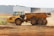 A large, yellow articulated dump truck is moving across a construction site. The truck has a spacious, open cargo area and oversized black tires. Dust is being kicked up by its movement, indicating it is actively being used in a dirt-laden environment. In the background, an industrial building with a flat roof stands near the site.