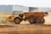 A large, yellow articulated dump truck is moving across a construction site. The truck has a spacious, open cargo area and oversized black tires. Dust is being kicked up by its movement, indicating it is actively being used in a dirt-laden environment. In the background, an industrial building with a flat roof stands near the site.