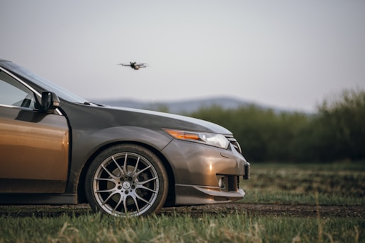 A close-up view of the front of a car parked on grass, with a drone flying in the background. The car is metallic gray in color, featuring sporty wheels and a sleek design. The landscape behind shows a blurred view of low hills and greenery.
