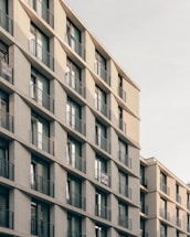 A multi-story residential building featuring repetitive balconies and windows with railings. The structure is made of light-colored bricks and has a modern architectural design. The perspective is angled, showing the edges and depth of the building.