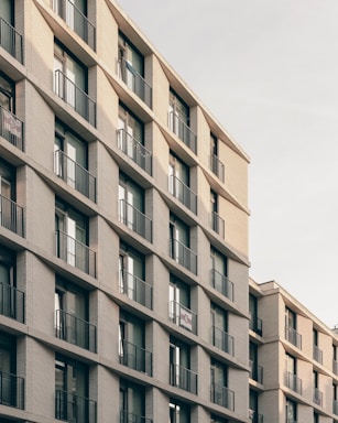 A multi-story residential building featuring repetitive balconies and windows with railings. The structure is made of light-colored bricks and has a modern architectural design. The perspective is angled, showing the edges and depth of the building.