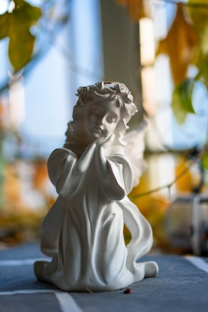 A serene porcelain angel figurine with a peaceful expression sits gracefully with hands under its chin. The sculpture has delicate facial features, flowing robes, and small wings. The background is softly blurred with hints of natural light and foliage, creating a tranquil atmosphere.