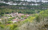 A quaint village nestled in a valley, surrounded by lush greenery and a backdrop of rocky cliffs. The village consists of rustic houses with brown and red rooftops, clustered around a tall, central building that appears to be a church. Vibrant green trees and fields dominate the landscape, creating a serene and peaceful atmosphere.