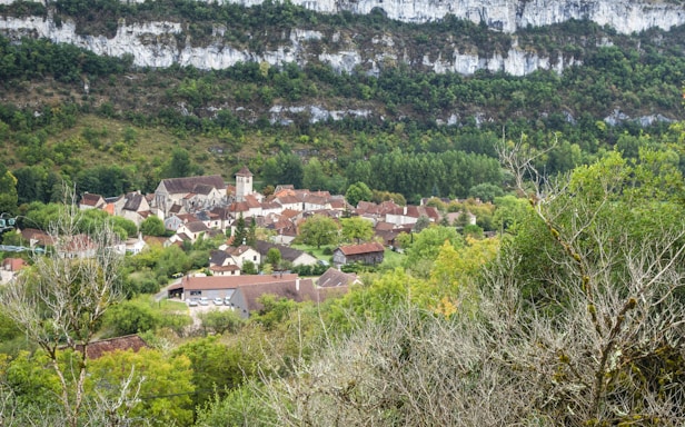A quaint village nestled in a valley, surrounded by lush greenery and a backdrop of rocky cliffs. The village consists of rustic houses with brown and red rooftops, clustered around a tall, central building that appears to be a church. Vibrant green trees and fields dominate the landscape, creating a serene and peaceful atmosphere.