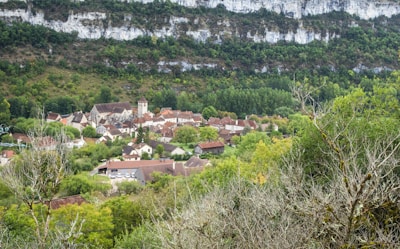 A quaint village nestled in a valley, surrounded by lush greenery and a backdrop of rocky cliffs. The village consists of rustic houses with brown and red rooftops, clustered around a tall, central building that appears to be a church. Vibrant green trees and fields dominate the landscape, creating a serene and peaceful atmosphere.