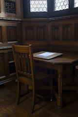 A small wooden desk and chair in a corner of a room with intricate wood paneling and two stained glass windows.