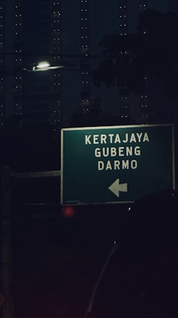 A directional road sign is illuminated in a dark urban setting, with the words 'Kertajaya,' 'Gubeng,' and 'Darmo' displayed, along with an arrow pointing left. The background features dimly lit high-rise buildings and a streetlamp casting light.