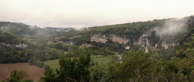 A misty valley with lush greenery and rocky cliffs. Trees and vegetation cover the landscape, with a few houses nestled among the hills. The cliffs are prominent and rugged, adding a dramatic backdrop to the serene and natural setting.