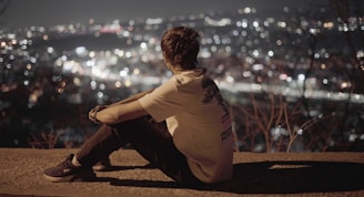 A person sits on a ledge, gazing out at a cityscape illuminated with numerous twinkling lights, suggesting an evening or nighttime setting. They appear to be deep in thought, with a serene and contemplative atmosphere surrounding them.