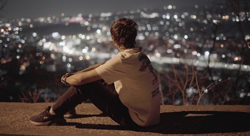 A person sits on a ledge, gazing out at a cityscape illuminated with numerous twinkling lights, suggesting an evening or nighttime setting. They appear to be deep in thought, with a serene and contemplative atmosphere surrounding them.