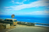 A tan van is parked beside a coastal road with a safety mirror. The scene overlooks the vast ocean under a partly cloudy sky, with the horizon stretching far in the distance. A concrete wall separates the road from the cliff edge providing a sense of safety.