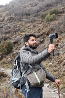 A man is hiking in a mountainous area while holding a camera mounted on a tripod for recording. He is wearing a gray puffer jacket and carrying a large backpack and a waist pouch. The terrain is rocky with sparse vegetation, and there is a walking stick in his other hand.