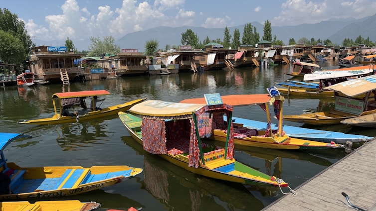 Numerous traditional boats with colorful canopies float on a serene water body, with an array of houseboats lined up in the background. The sky is clear with scattered clouds, and there are lush green trees bordering the area. Mountains are visible in the distance.