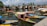 Numerous traditional boats with colorful canopies float on a serene water body, with an array of houseboats lined up in the background. The sky is clear with scattered clouds, and there are lush green trees bordering the area. Mountains are visible in the distance.