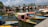 Numerous traditional boats with colorful canopies float on a serene water body, with an array of houseboats lined up in the background. The sky is clear with scattered clouds, and there are lush green trees bordering the area. Mountains are visible in the distance.