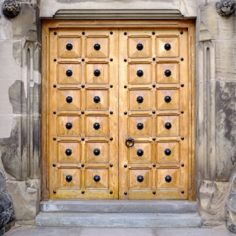 A large wooden door with intricate carvings and black metal studs arranged in a grid pattern. The door is surrounded by a stone frame with ornate details and textures. The wood has a warm, golden tone and the stone features weathered and varied shades of gray.
