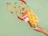 A hand with red nail polish holds a bag of nacho chips that appears to be open. Various nacho chips, red chili peppers, peppercorns, and nuts seem to be dynamically emerging from the bag. The background is a solid light green color.