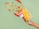 A hand with red nail polish holds a bag of nacho chips that appears to be open. Various nacho chips, red chili peppers, peppercorns, and nuts seem to be dynamically emerging from the bag. The background is a solid light green color.