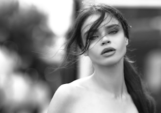 A black and white portrait of a woman with long hair and an expressive gaze. The wind blows strands of her hair across her face, adding a sense of motion and intimacy to the image. The background is softly blurred, focusing attention on her features.