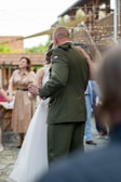A couple is dancing outdoors, with the man wearing a military uniform and the woman in a wedding dress. They are surrounded by softly lit string lights, creating a romantic atmosphere. Several people in the background are watching, contributing to a festive ambiance.