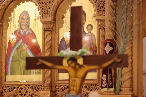Wooden crucifix with a figure of Jesus in the foreground, set against a richly decorated golden religious icon panel featuring two saints. The carvings are intricate, and soft lighting highlights the ornate woodwork and the colorful robes on the icons.