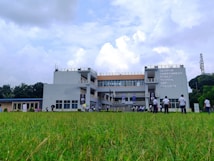 A multi-story educational institution is surrounded by a grassy field. Several people wearing uniforms are gathered in front of the building, some walking across the lawn. The sky is partly cloudy, and there is signage on the building indicating a school name.