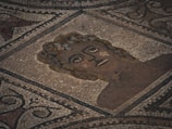 A mosaic artwork featuring a stylized human face surrounded by intricate geometric and swirling patterns, composed of small, colored tiles in earthy tones.