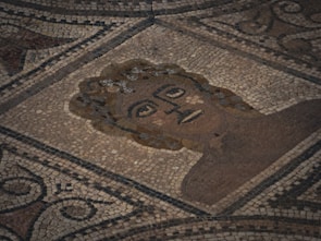 A mosaic artwork featuring a stylized human face surrounded by intricate geometric and swirling patterns, composed of small, colored tiles in earthy tones.