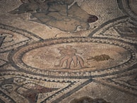 An ancient mosaic depicting two human figures in close proximity within an oval boundary, surrounded by intricate patterns and shapes that form a detailed artistic composition.