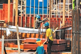 Workers wearing helmets and safety gear are engaged in construction activities on a building site with scaffolding and wooden and metal frames visible.