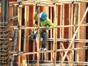 A construction worker wearing a blue hard hat and green safety vest works amidst bamboo scaffolding. The scene includes various construction materials such as wooden planks and metal reinforcements.