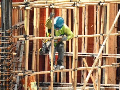 A construction worker wearing a blue hard hat and green safety vest works amidst bamboo scaffolding. The scene includes various construction materials such as wooden planks and metal reinforcements.