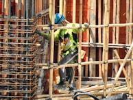 A construction worker wearing a blue helmet and green safety vest is carefully navigating through a framework of wooden beams. The worker is positioned within a partially built structure, which includes vertical and horizontal wooden and metal bars. The scene is busy with various construction materials visible.