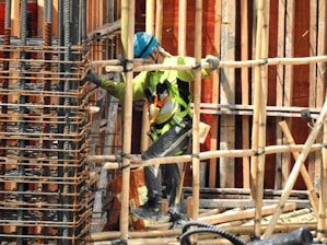 A construction worker wearing a blue helmet and green safety vest is carefully navigating through a framework of wooden beams. The worker is positioned within a partially built structure, which includes vertical and horizontal wooden and metal bars. The scene is busy with various construction materials visible.