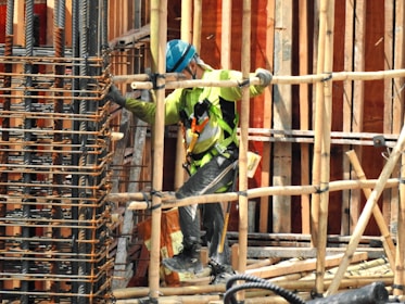 A construction worker wearing a blue helmet and green safety vest is carefully navigating through a framework of wooden beams. The worker is positioned within a partially built structure, which includes vertical and horizontal wooden and metal bars. The scene is busy with various construction materials visible.