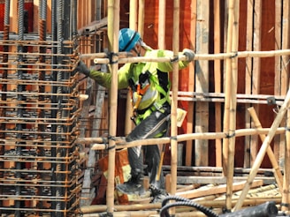 A construction worker wearing a blue helmet and green safety vest is carefully navigating through a framework of wooden beams. The worker is positioned within a partially built structure, which includes vertical and horizontal wooden and metal bars. The scene is busy with various construction materials visible.