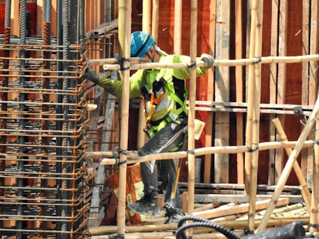 A construction worker wearing a blue helmet and green safety vest is carefully navigating through a framework of wooden beams. The worker is positioned within a partially built structure, which includes vertical and horizontal wooden and metal bars. The scene is busy with various construction materials visible.