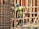 A construction worker wearing a blue helmet and green safety vest is carefully navigating through a framework of wooden beams. The worker is positioned within a partially built structure, which includes vertical and horizontal wooden and metal bars. The scene is busy with various construction materials visible.