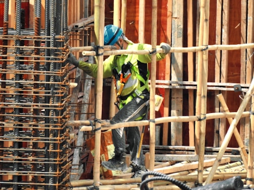 A construction worker wearing a blue helmet and green safety vest is carefully navigating through a framework of wooden beams. The worker is positioned within a partially built structure, which includes vertical and horizontal wooden and metal bars. The scene is busy with various construction materials visible.