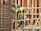 A construction worker wearing a blue helmet and green safety vest is carefully navigating through a framework of wooden beams. The worker is positioned within a partially built structure, which includes vertical and horizontal wooden and metal bars. The scene is busy with various construction materials visible.