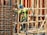 A construction worker wearing a blue helmet and green safety vest is carefully navigating through a framework of wooden beams. The worker is positioned within a partially built structure, which includes vertical and horizontal wooden and metal bars. The scene is busy with various construction materials visible.