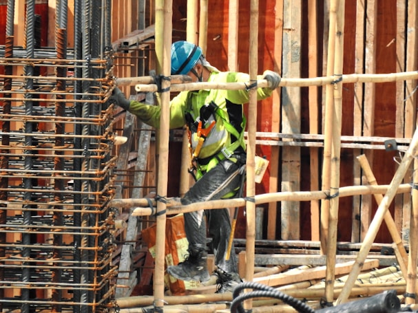 A construction worker wearing a blue helmet and green safety vest is carefully navigating through a framework of wooden beams. The worker is positioned within a partially built structure, which includes vertical and horizontal wooden and metal bars. The scene is busy with various construction materials visible.