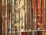 Numerous vertical metal rods, likely steel rebar, positioned closely together. Intermixed with these rods are bamboo poles, lashed together, perhaps for scaffolding or a construction project. The scene appears dense and intricate, with the warm, rust-like tones of the metal contrasting against the paler bamboo.