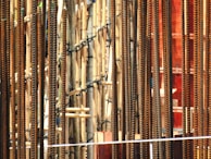 Numerous vertical metal rods, likely steel rebar, positioned closely together. Intermixed with these rods are bamboo poles, lashed together, perhaps for scaffolding or a construction project. The scene appears dense and intricate, with the warm, rust-like tones of the metal contrasting against the paler bamboo.