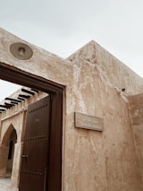 A traditional building with textured beige stone walls and a solid wooden door. A sign mounted on the wall reads 'Utensils Market' in both English and Arabic. The architecture features simple geometric shapes and an open roof structure with visible beams.
