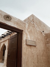 A traditional building with textured beige stone walls and a solid wooden door. A sign mounted on the wall reads 'Utensils Market' in both English and Arabic. The architecture features simple geometric shapes and an open roof structure with visible beams.
